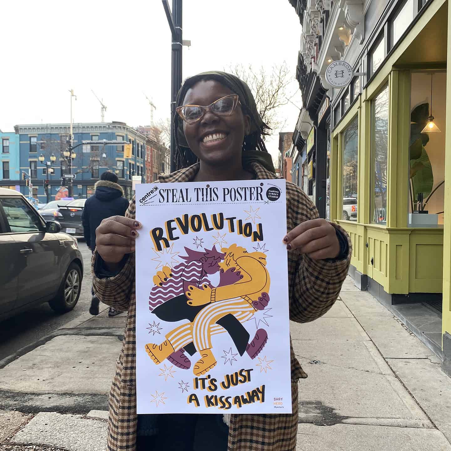 Smiling person holding a poster that reads “Revolution – It’s Just a Kiss Away” on a city sidewalk with storefronts and traffic in the background