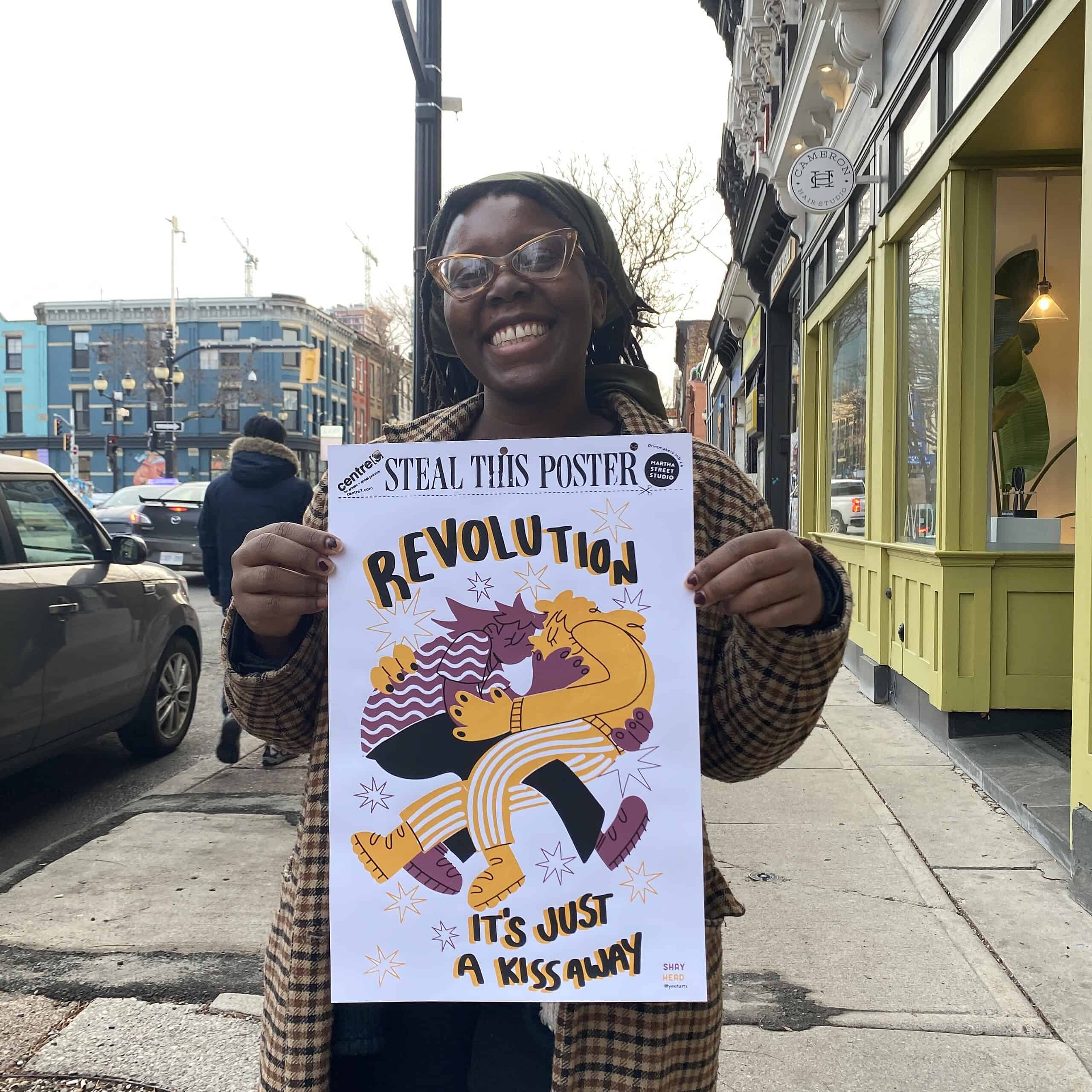 Smiling person holding a poster that reads “Revolution – It’s Just a Kiss Away” on a city sidewalk with storefronts and traffic in the background