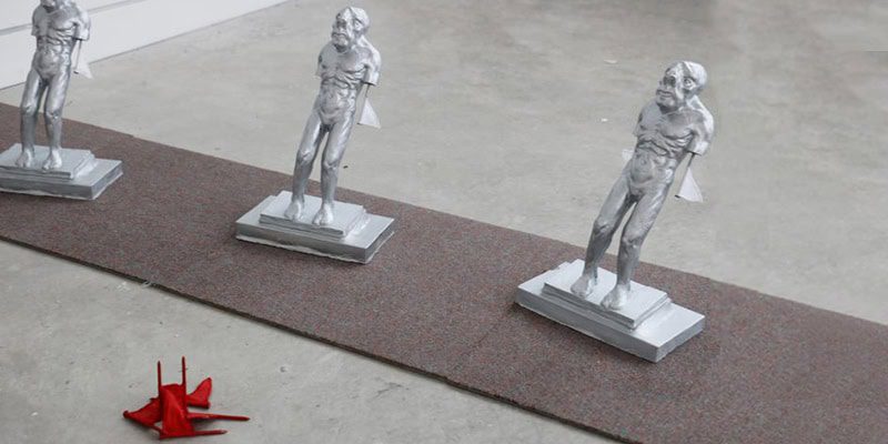 Small silver figurative sculptures arranged in a line on a carpeted platform with a red object placed nearby