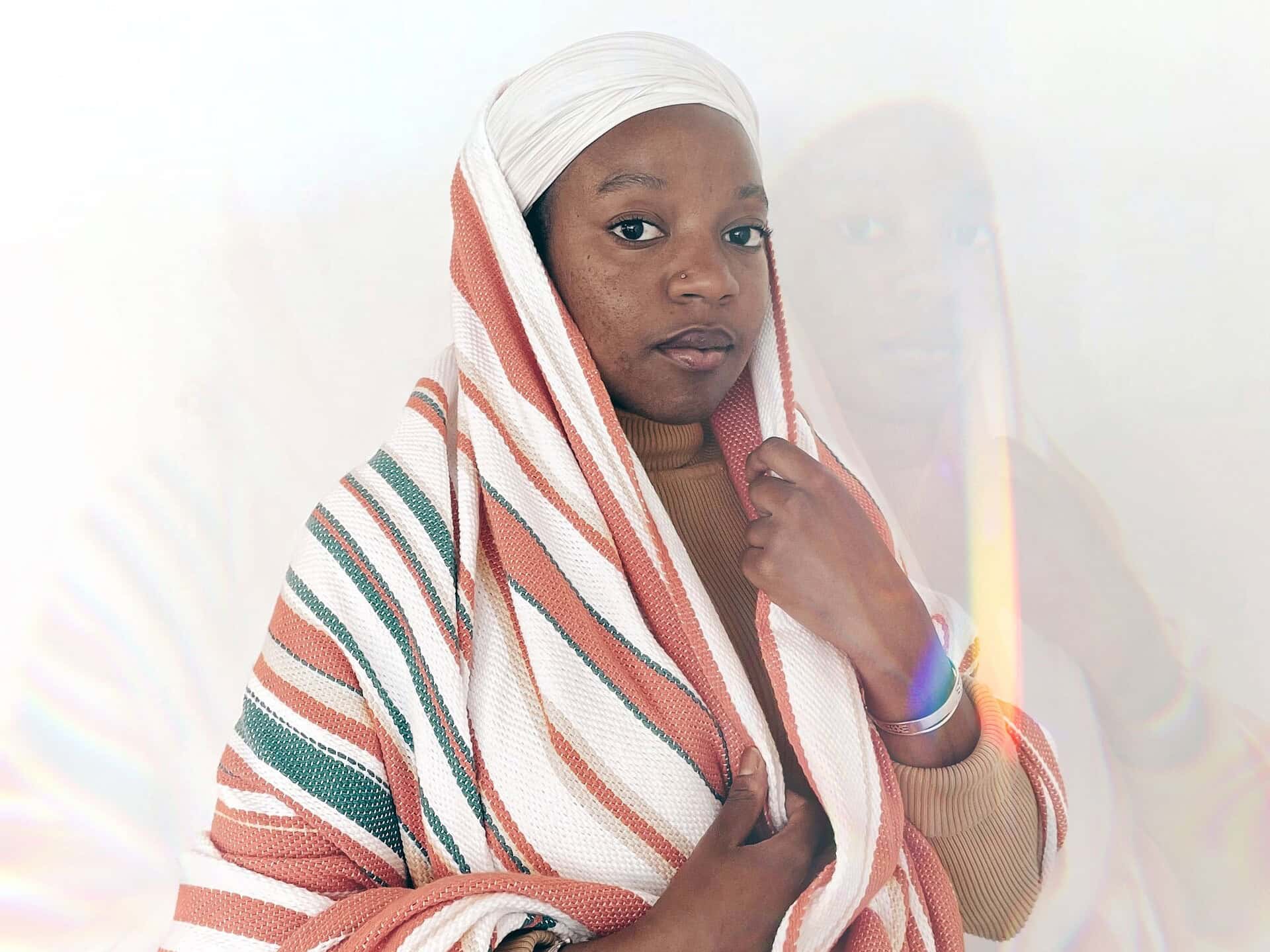 Portrait of a person wearing a striped woven textile draped over her head and shoulders, holding the fabric close; soft lighting creates a faint double exposure effect against a pale background
