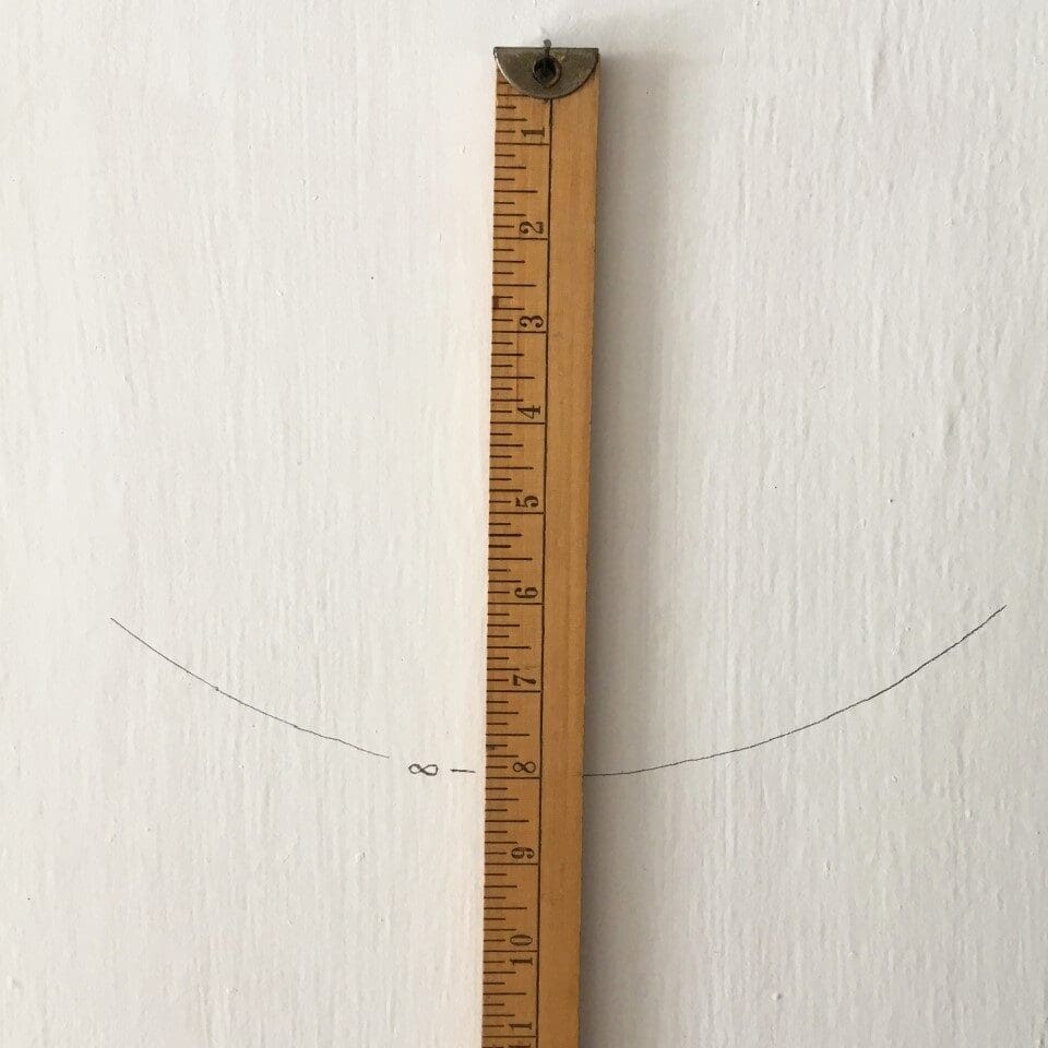 Wooden measuring stick mounted vertically on a white wall with a faint curved pencil line drawn across it