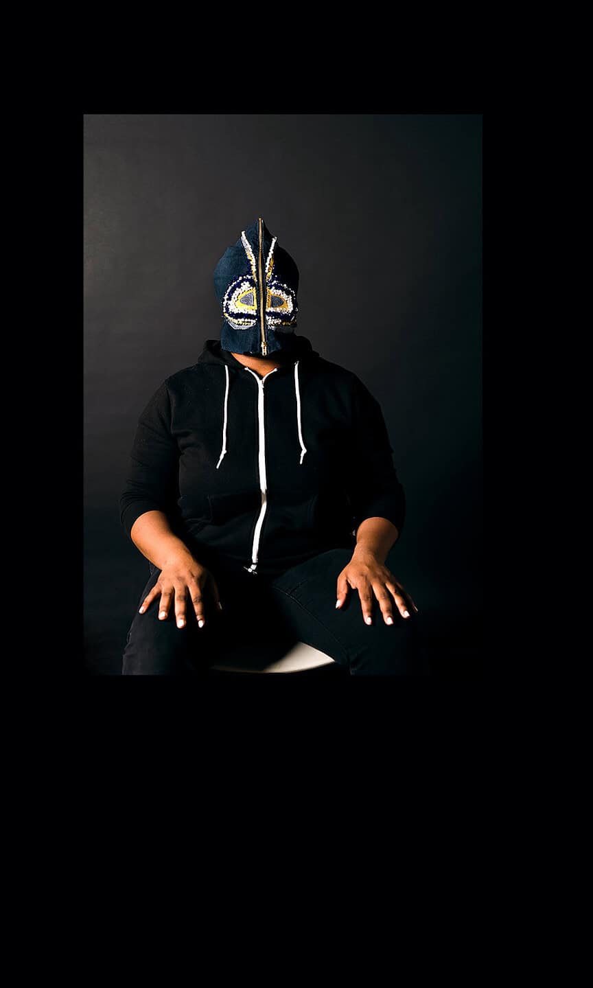 Seated figure wearing a black hoodie with a beaded mask featuring symmetrical patterns and a central zipper