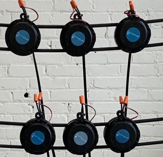 Six round black electronic devices with orange connectors are attached to black cables, arranged in two rows of three, in front of a white brick wall.