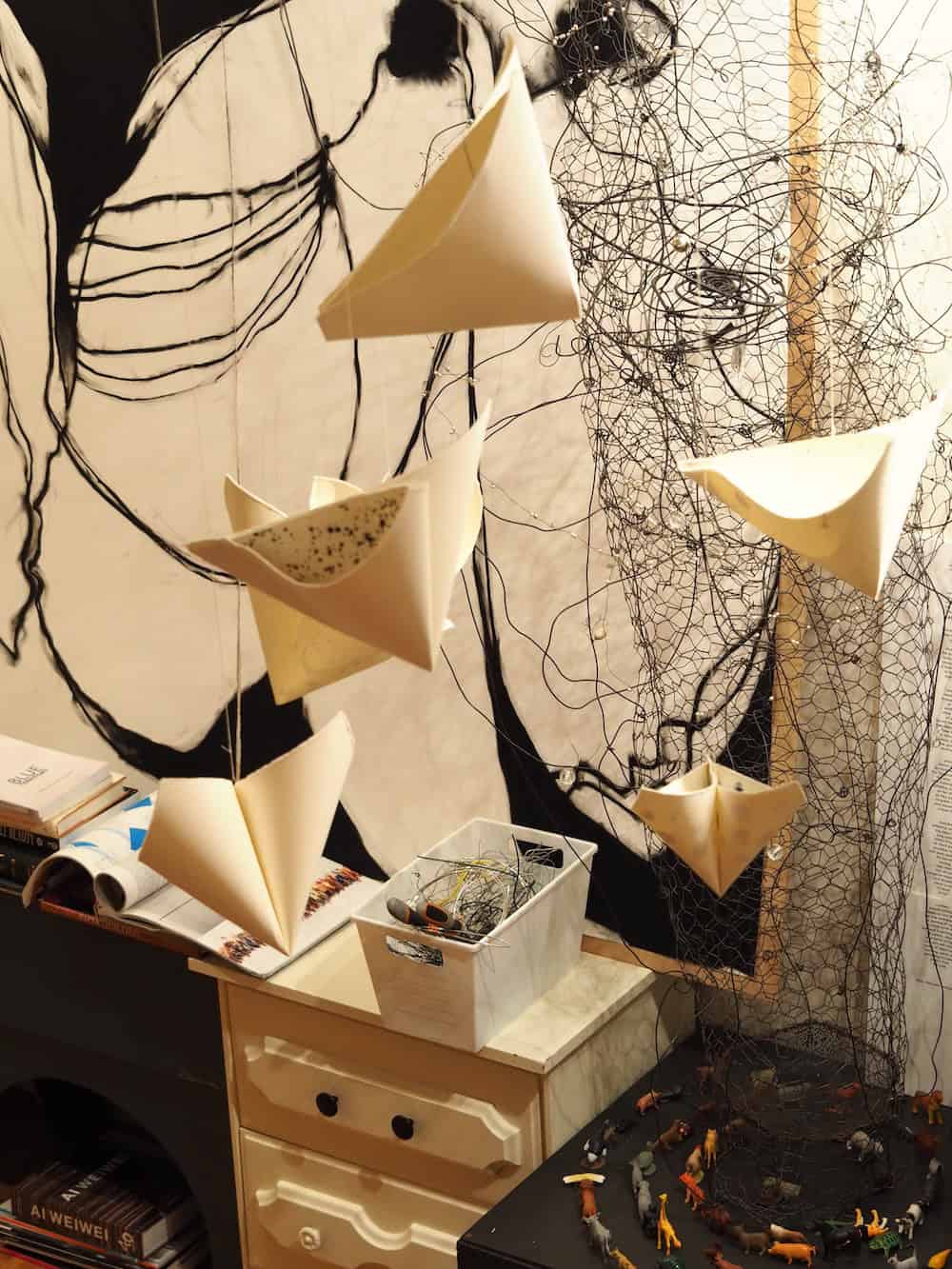 An installation view showing folded sculptural paper forms suspended in front of a net structure and expressive black line drawings on the wall.