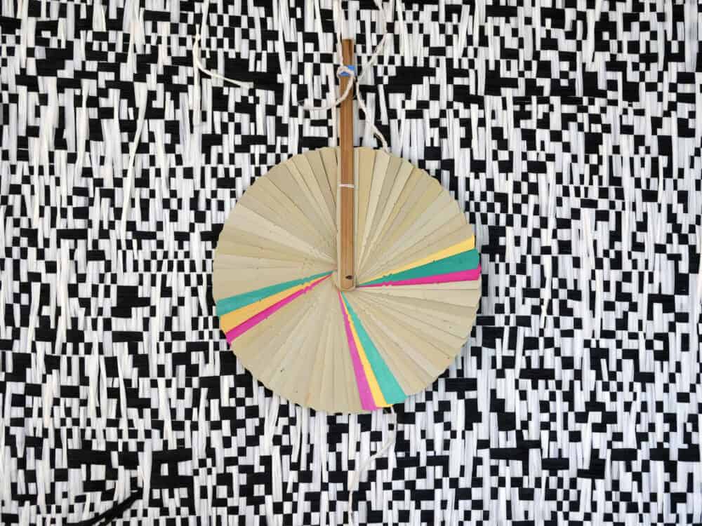 Close-up of a circular fan-like disc with coloured segments (green, pink, yellow) hanging in front of a black-and-white woven textile with a dense geometric pattern