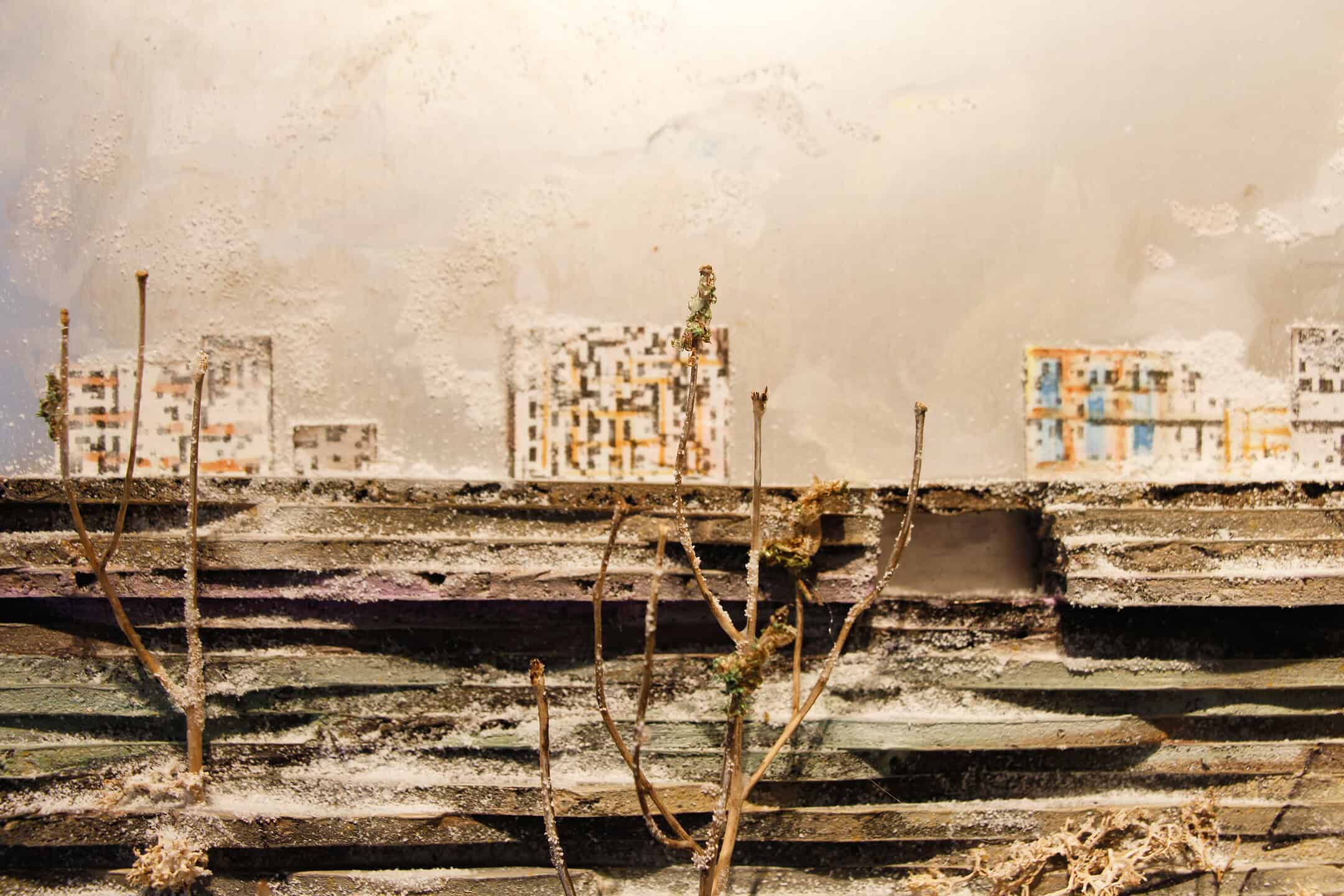 A close-up of a mixed-media landscape showing miniature painted building forms on a pale background, with twigs and textured material in the foreground.