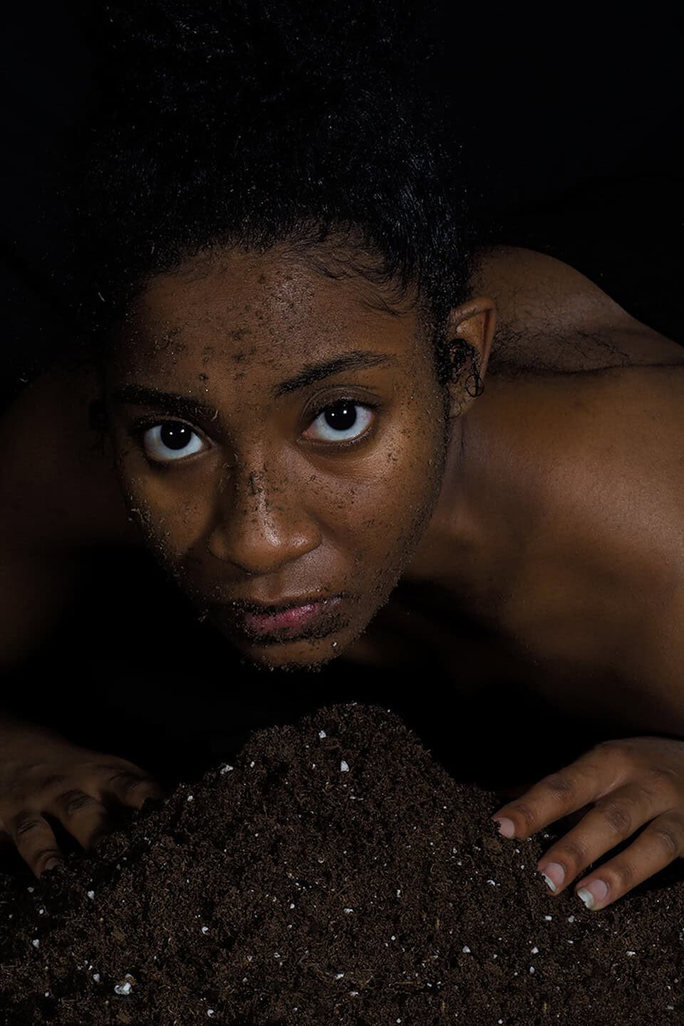 Close-up portrait of a person leaning over soil with dirt on their face, looking directly toward the viewer