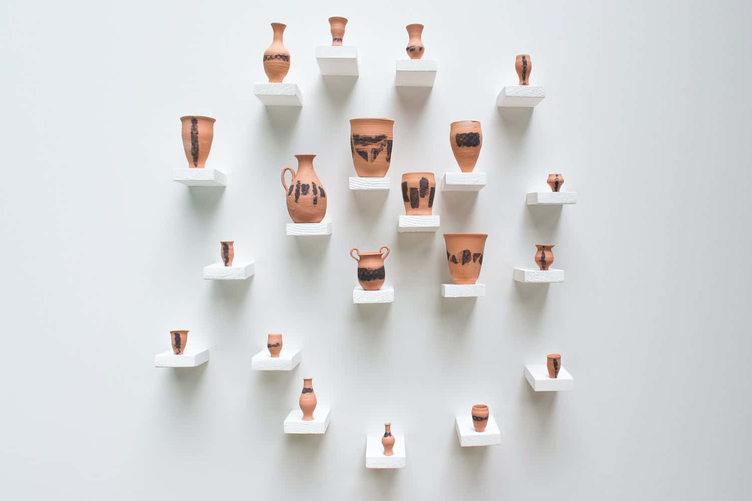 A wall installation of terracotta vessels arranged on staggered white shelves, each pot featuring unique black stamped patterns and organic shapes against a white gallery wall.