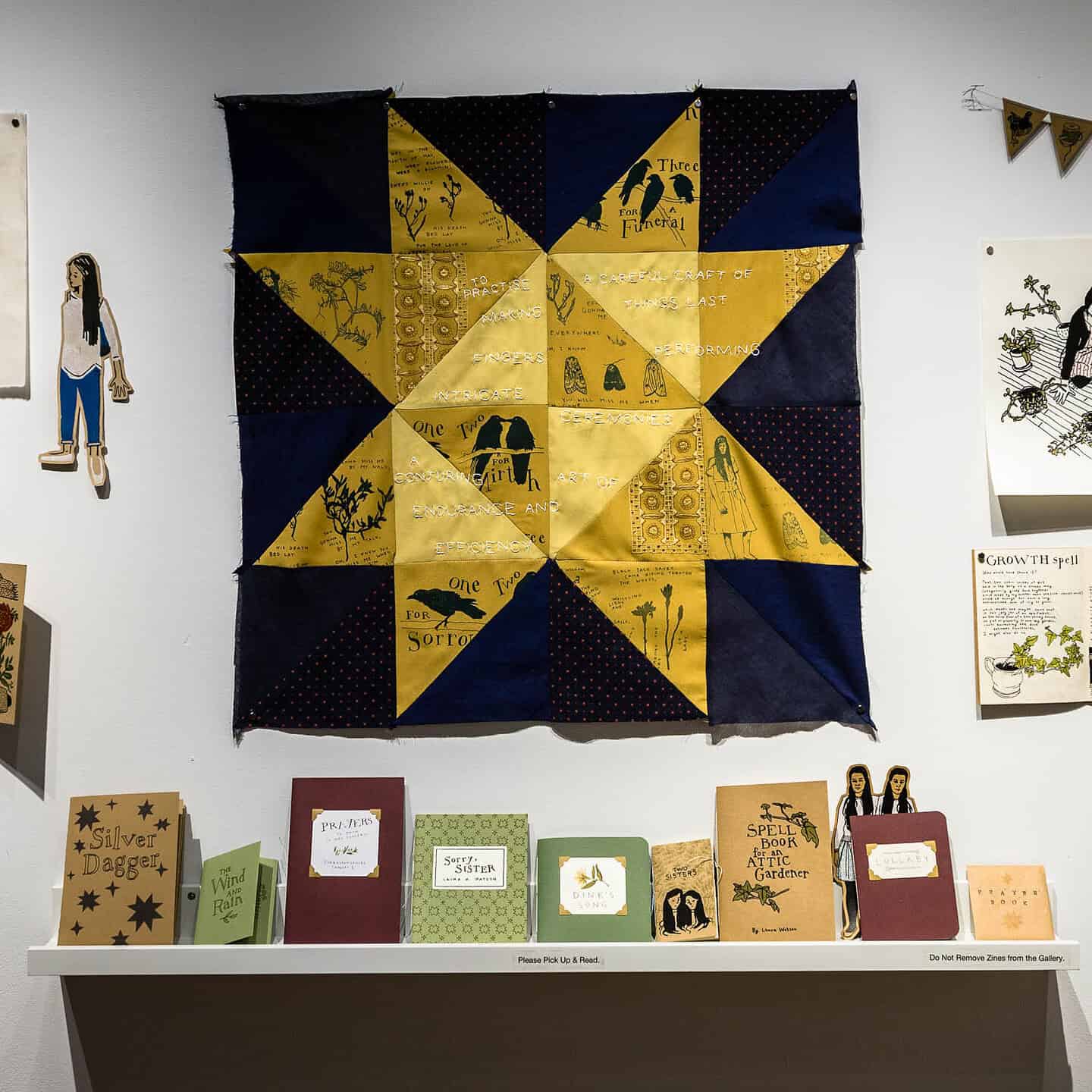 A close-up of an art display featuring a navy and yellow star-shaped quilt with printed text and bird illustrations, positioned above a shelf of colourful handmade zines.