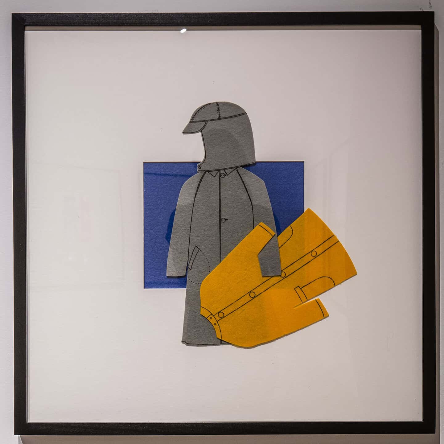 Framed felt-paper collage of a grey raincoat and matching cap, paired with an oversized yellow coat, set against a blue square background.