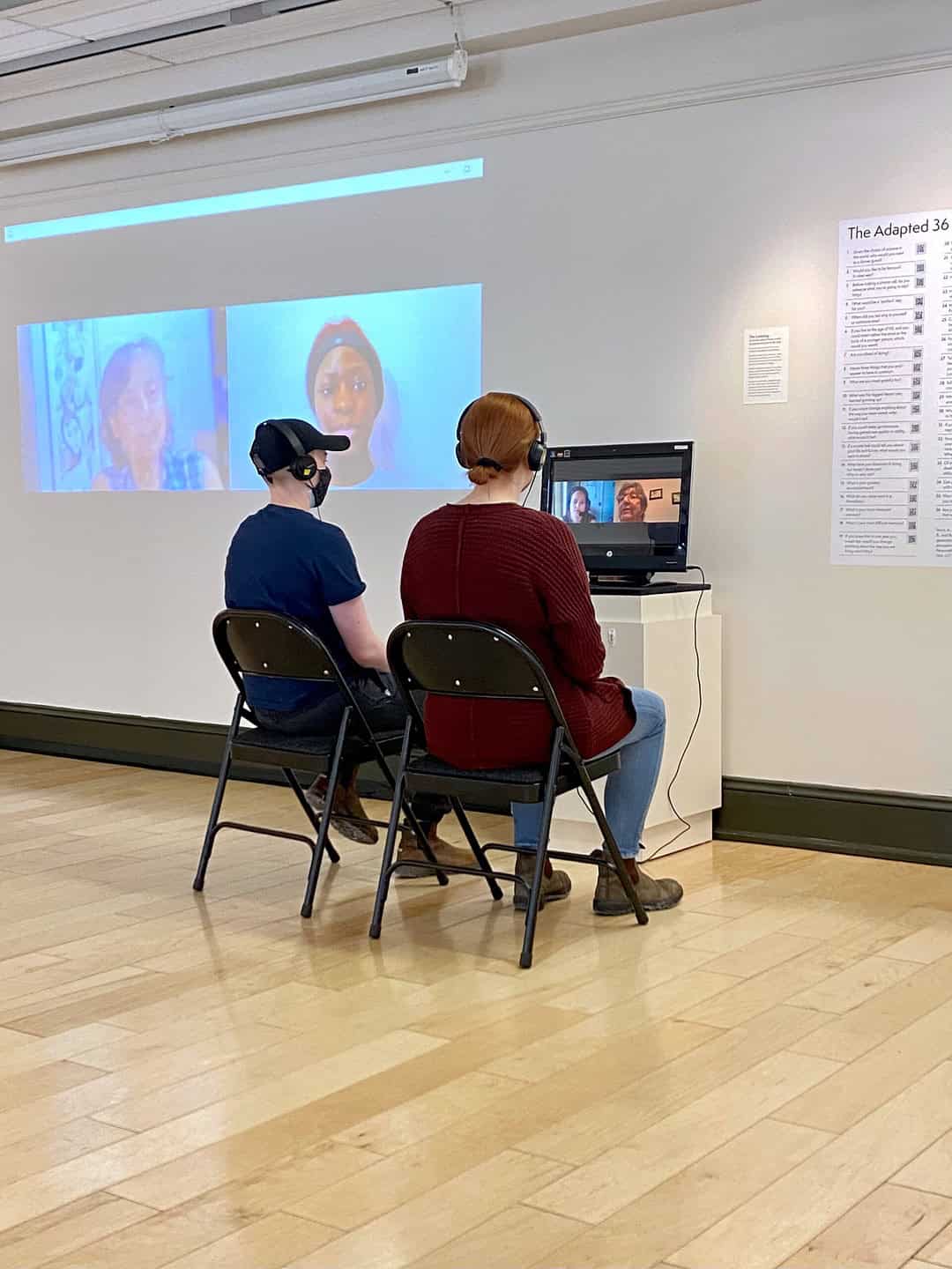 Two people wearing headphones sit on black chairs, facing a small TV screen mounted on a white stand. The TV shows a video call with two people smiling. The room has a wooden floor and a projection screen on the wall in front of them showing two more people on the call.