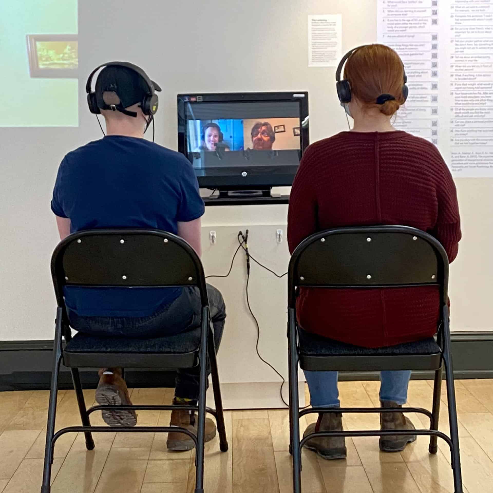 Two people wearing headphones sit facing a small screen showing a video call with two smiling participants.
