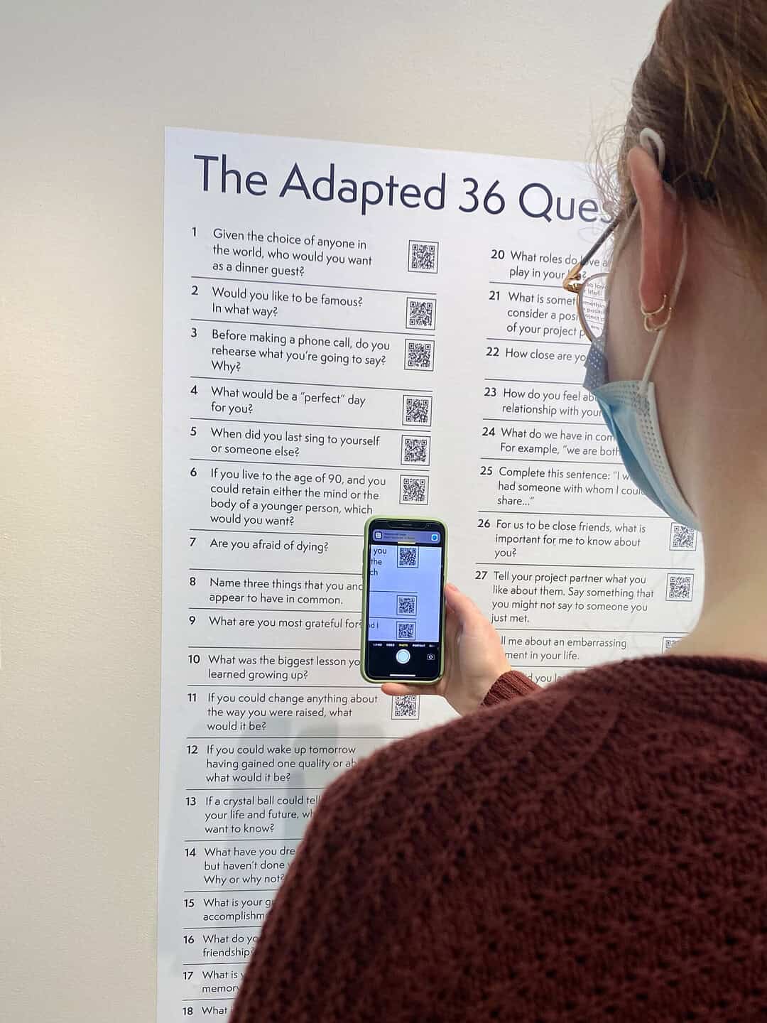 A person wearing glasses and a face mask scans a QR code on a printed Adapted 36 Questions poster using their smartphone.