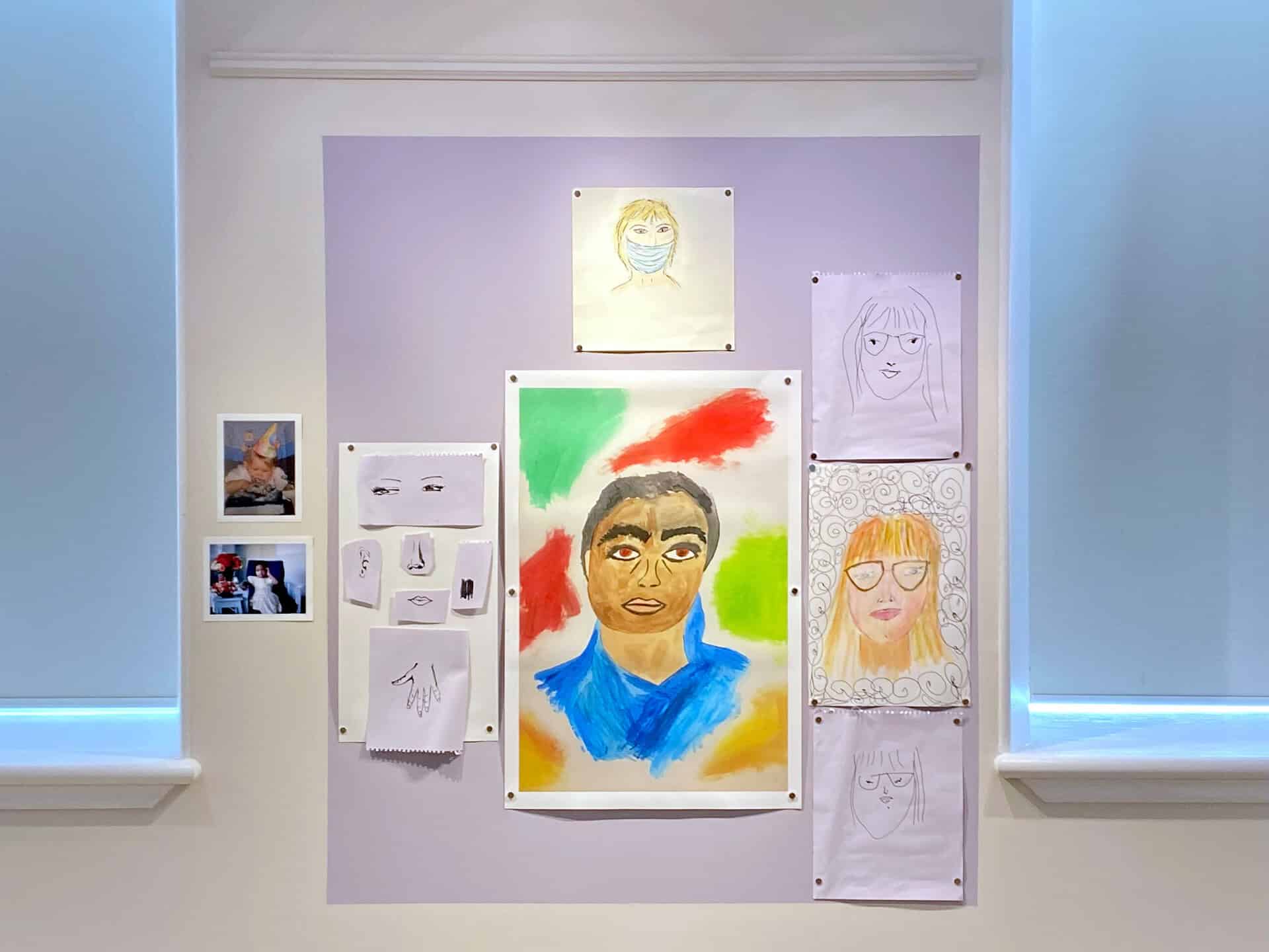 A wall display of art including a vibrant central portrait surrounded by pencil sketches of faces and hands, with two small photographs on the left, framed by two windows with white shades.