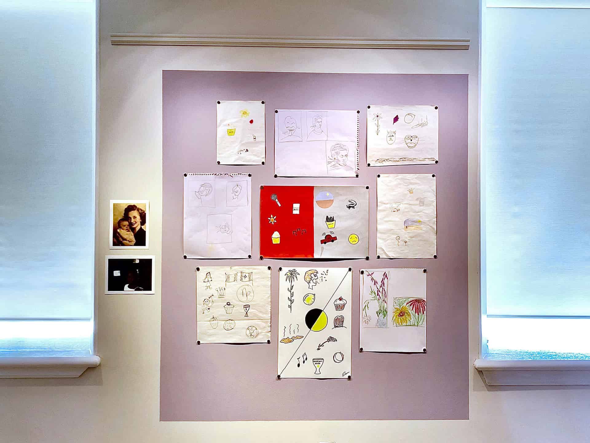 A wall display of twelve hand-drawn sketches and drawings is arranged in a grid, with two small printed photos on the left, framed by two windows with white shades.