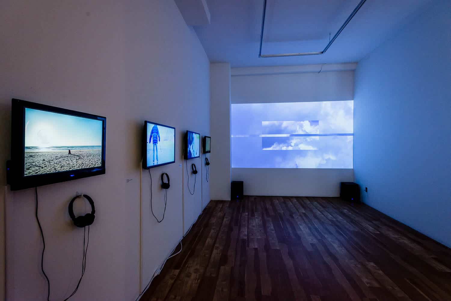 A side-angle perspective of the video installation. The monitors show various cinematic scenes, including a beach and a figure in a blue shirt. The large projection in the background displays a sky filled with soft white clouds.