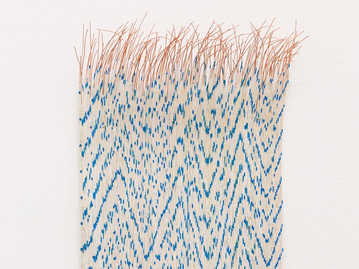 Close-up of a woven textile artwork with a zigzag and dotted blue pattern on a white background, with loose, frayed orange threads at the top and bottom edges, hanging against a white wall.