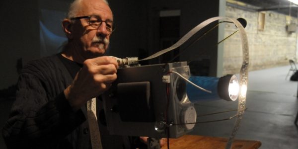 David Bobier, an individual with glasses and grey hair, carefully adjusts a mechanical sound or film device. A long strip of perforated paper tape loops through the machine, which emits a bright light from a blue cylindrical lens.