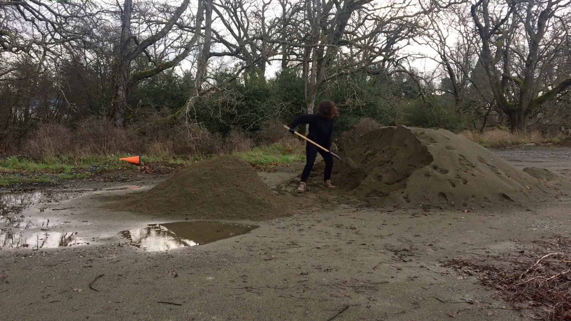 A person shovelling gravel from a large pile in an outdoor area with bare trees and puddles