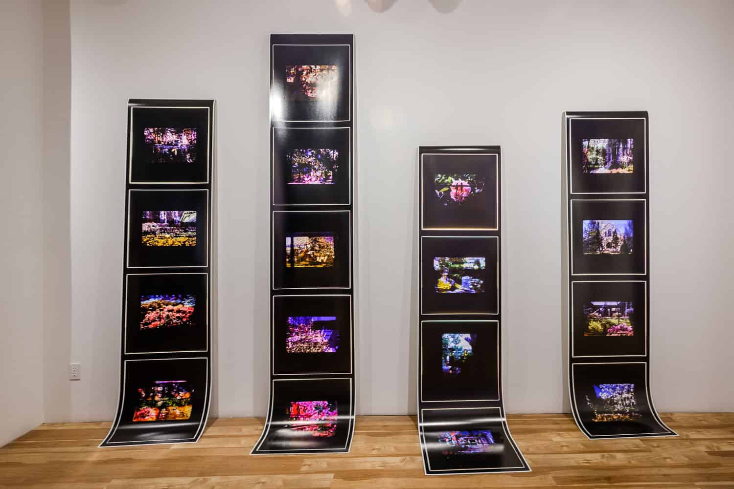 Three tall, vertical black panels each display four colourful, illuminated film slide photographs. The panels lean against a white wall and their lower sections curve onto a wooden floor.