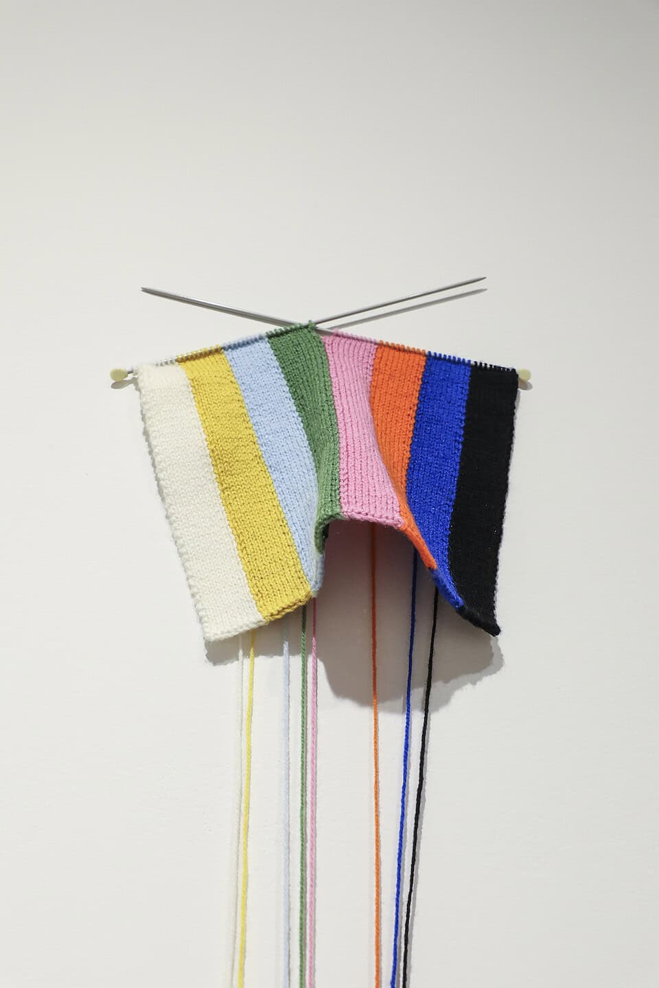 Small striped knitted panel pinned to the wall on crossed knitting needles, with vertical bands of cream, yellow, pale blue, green, pink, orange, blue, and black and loose strands of matching yarn hanging down beneath it