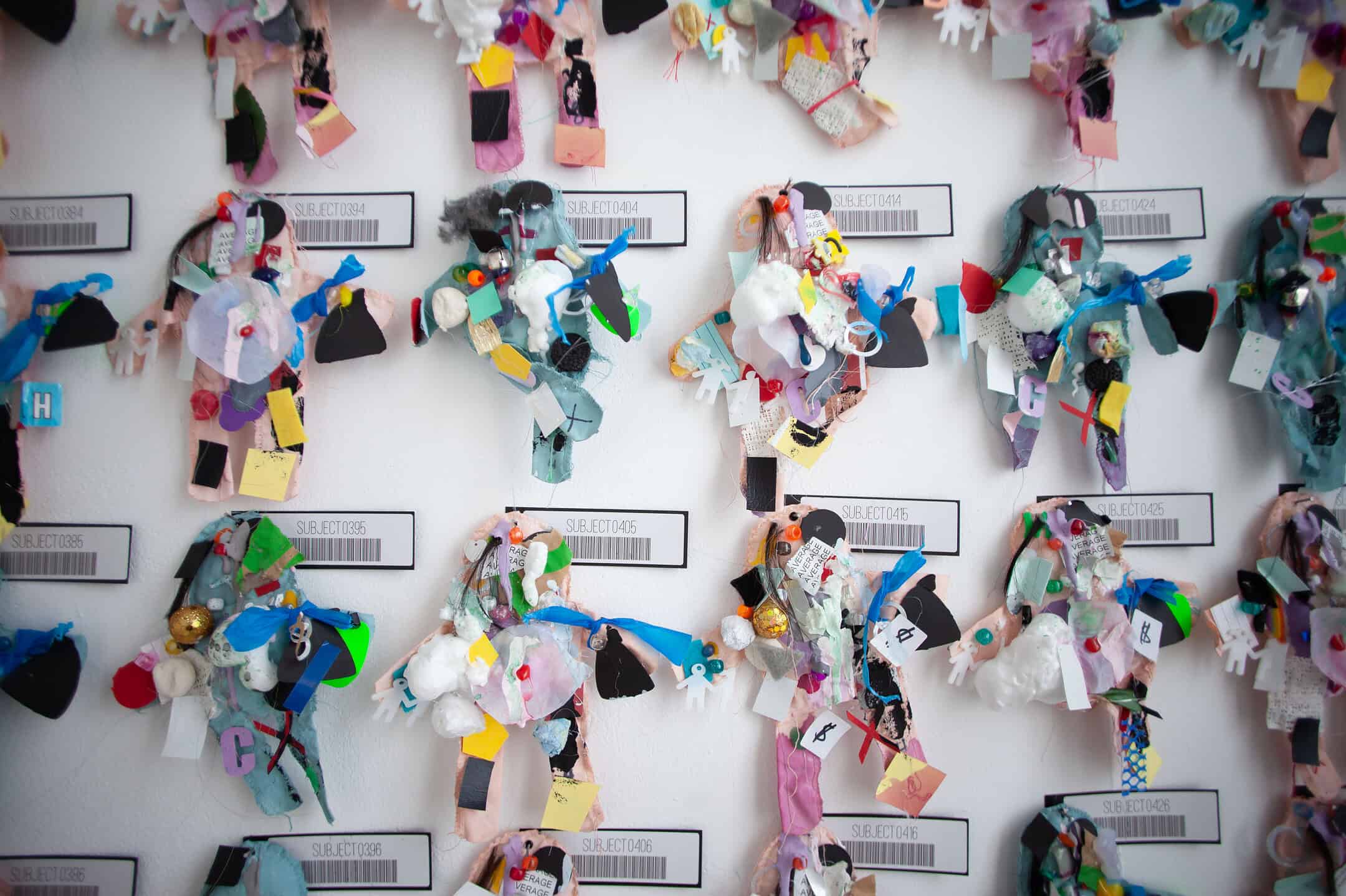 Abstract mixed-media art pieces made of colourful scraps, foam, and various materials are mounted on a wall above barcode labels reading SUBJECT0235 and similar variants.