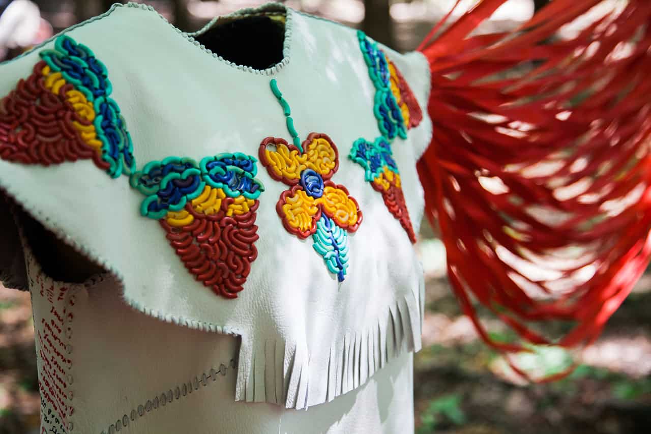 Close-up of a garment with bright floral beadwork and long red strands extending from the back against an outdoor background