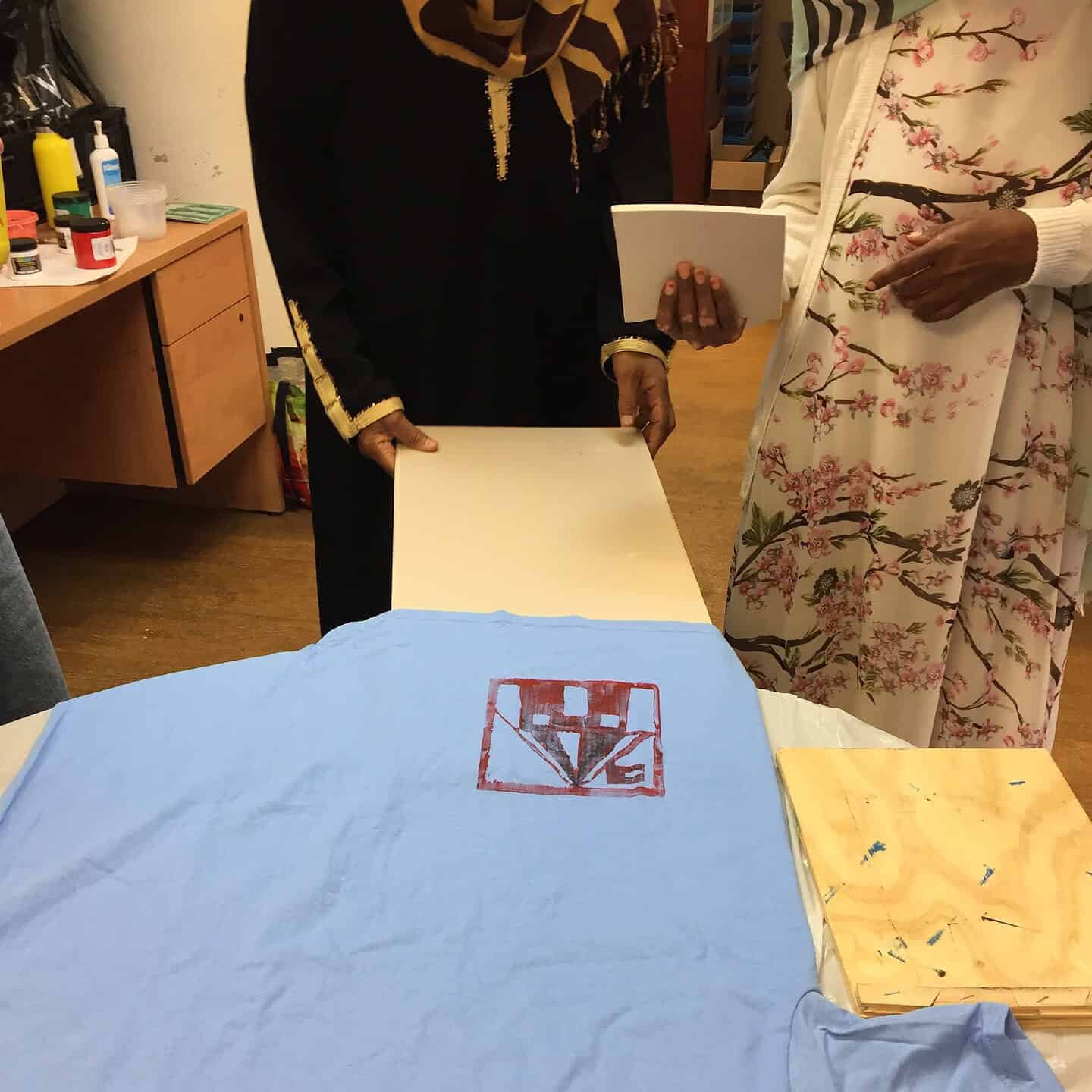 Two people stand at a table. One holds a wooden board, the other a white card. On the table lies a blue T-shirt with a red geometric print and some wooden blocks with paint stains nearby.