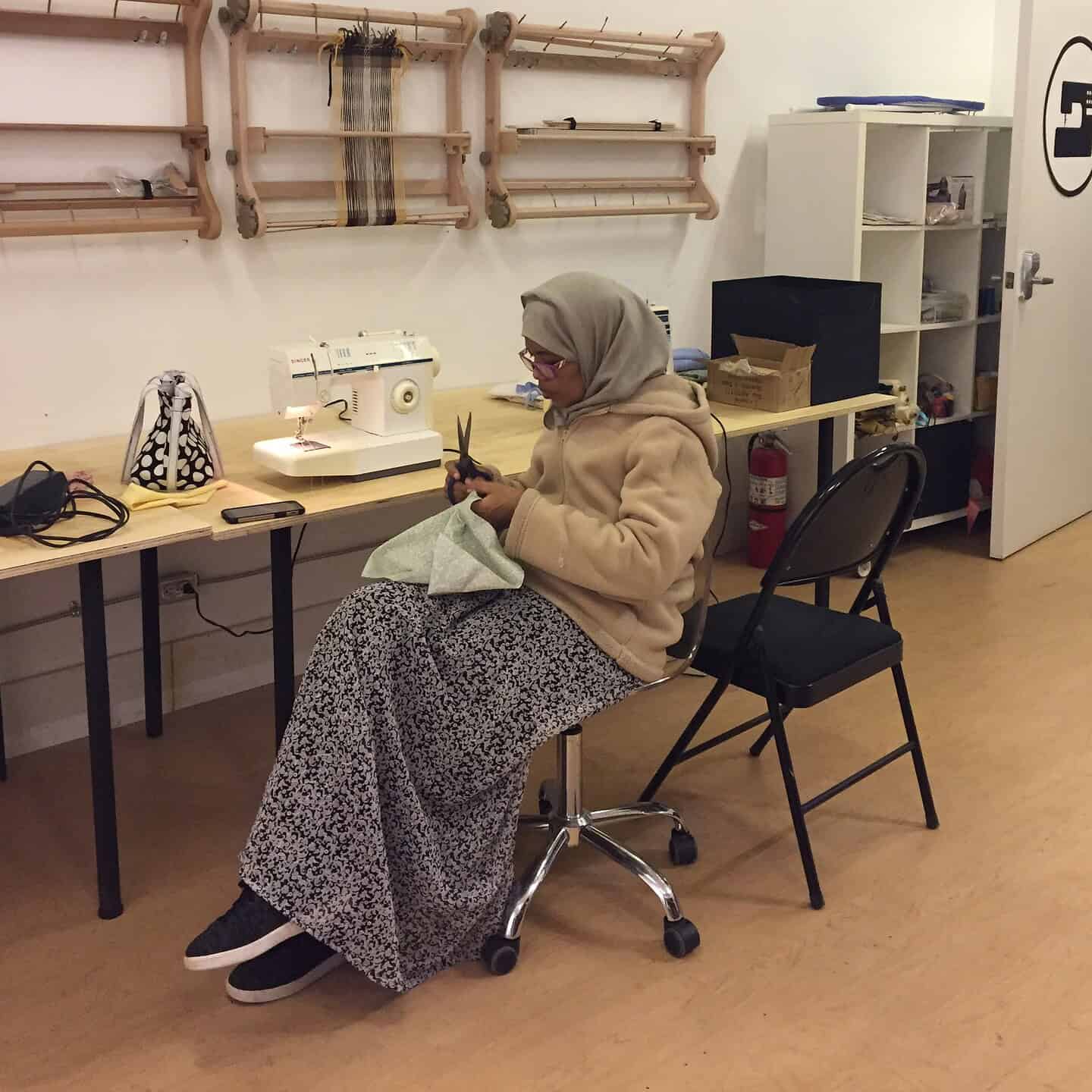 A person in a hijab and patterned skirt sits on a chair, cutting fabric. A sewing machine, shelves with supplies, and small hanging looms are visible in the room.