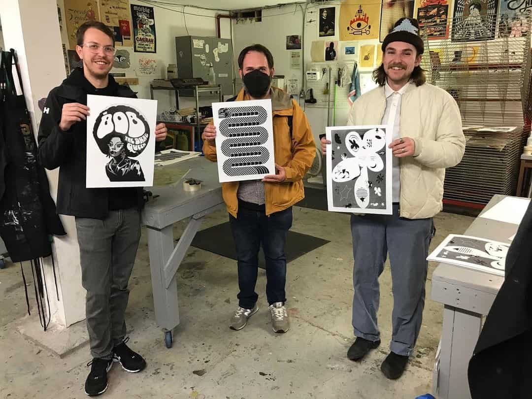 Three people stand in Centre[4]Arts' Screen Printing Studio, each holding a unique black-and-white print. Art supplies and colourful posters fill the background. The person in the centre wears a mask and a yellow jacket.