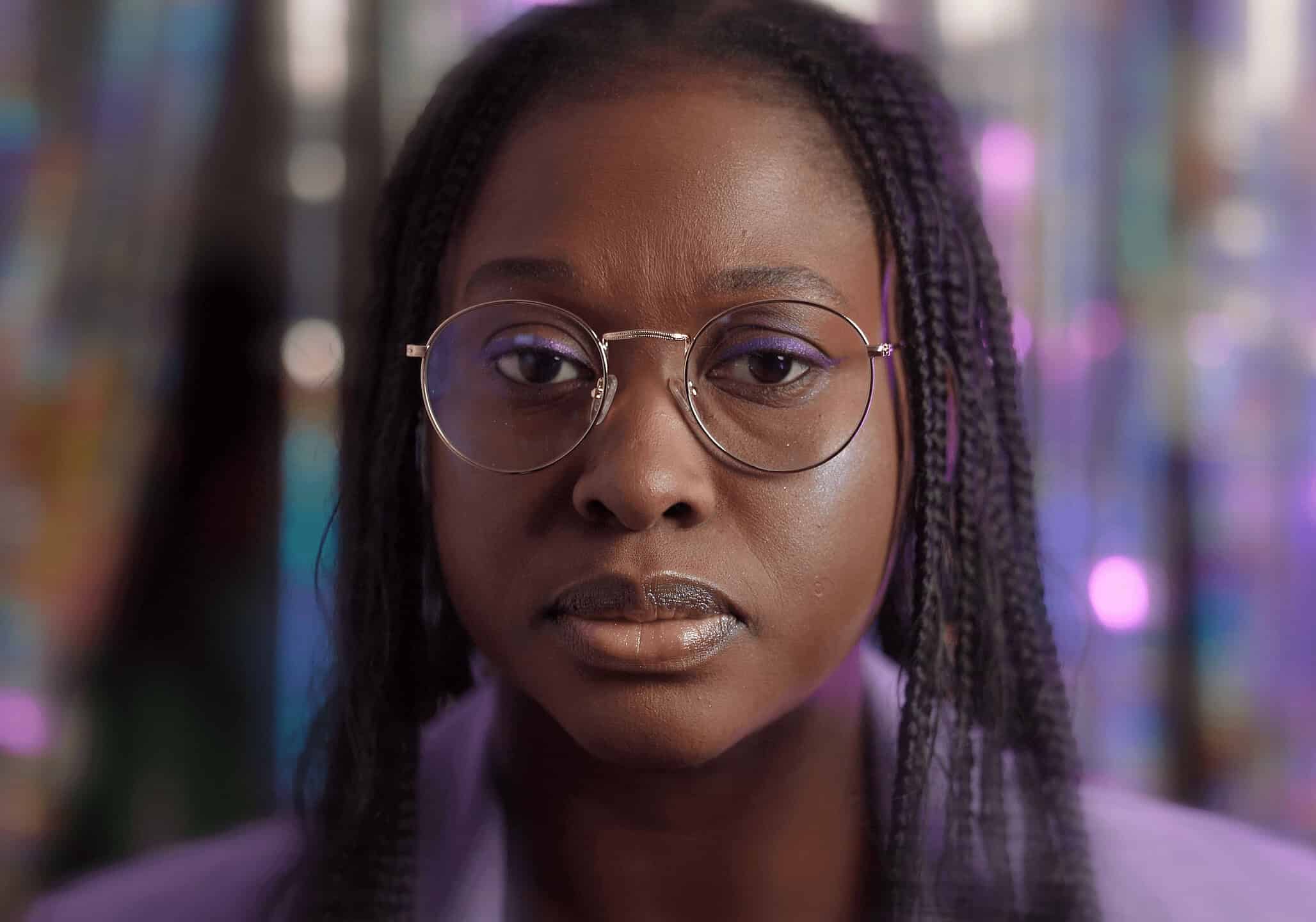A close-up cinematic portrait of a person with braided hair and round glasses. They have a serious expression, and the background is a soft-focus blur of shimmering, iridescent purple and blue light.