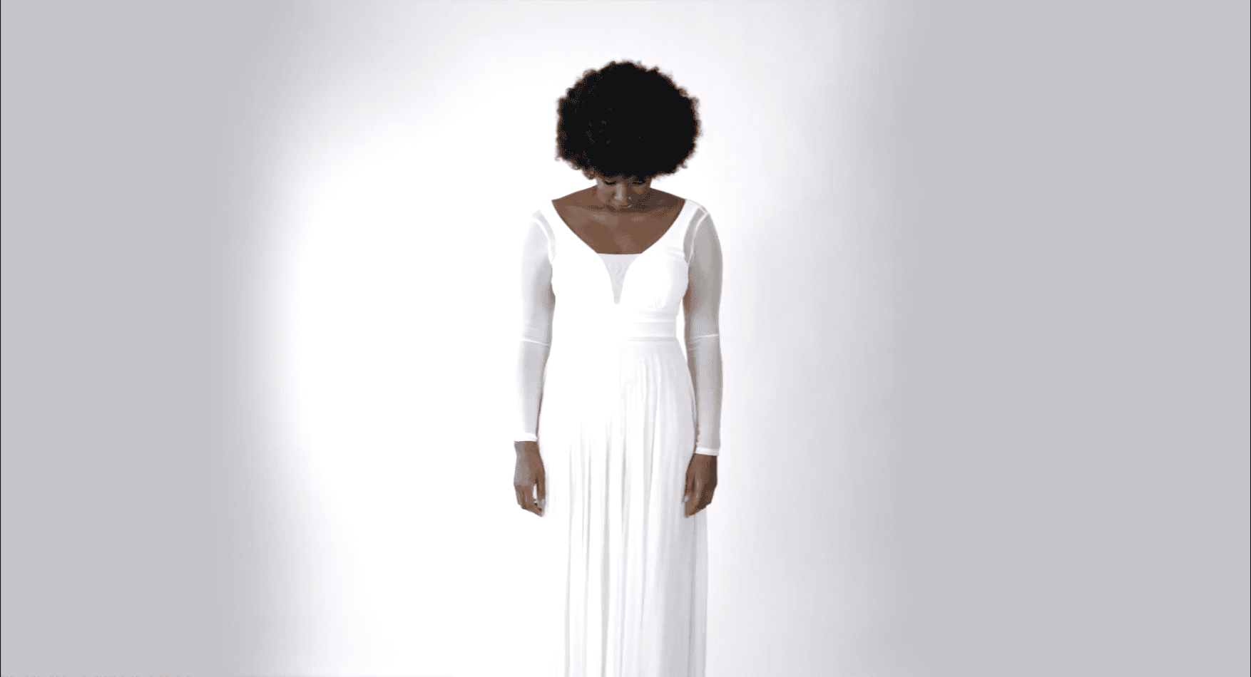 A still from a video work titled "ANGST." A person with an afro, wearing a long-sleeved white dress, stands against a bright white background with their head bowed in a contemplative or somber pose.
