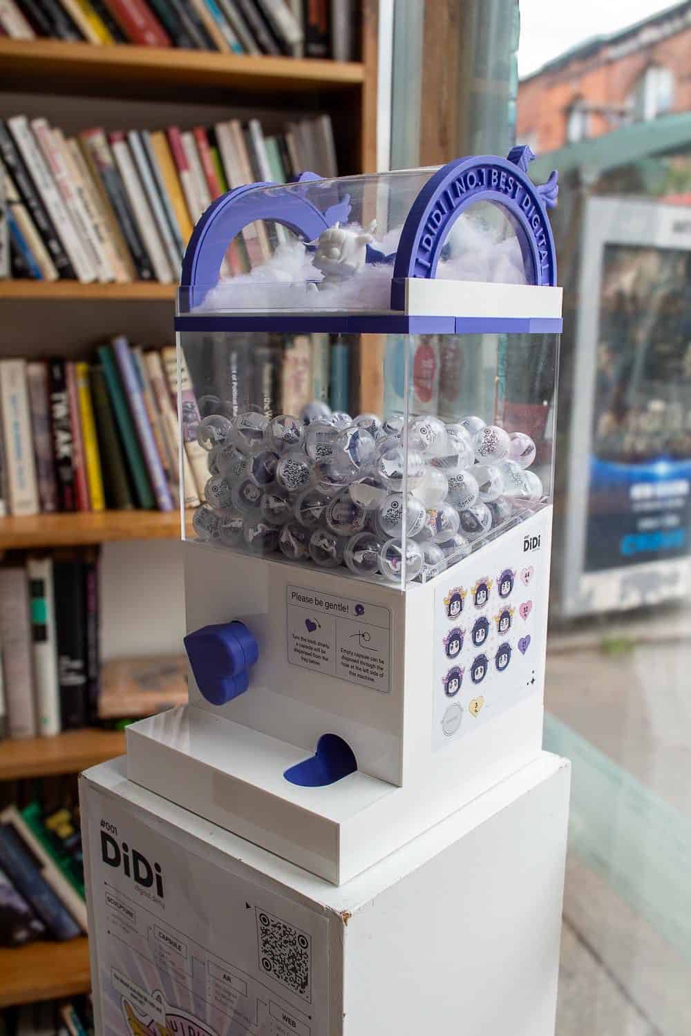 A white gacha machine sculpture filled with capsules, sitting on a plinth in front of a bookshelf.