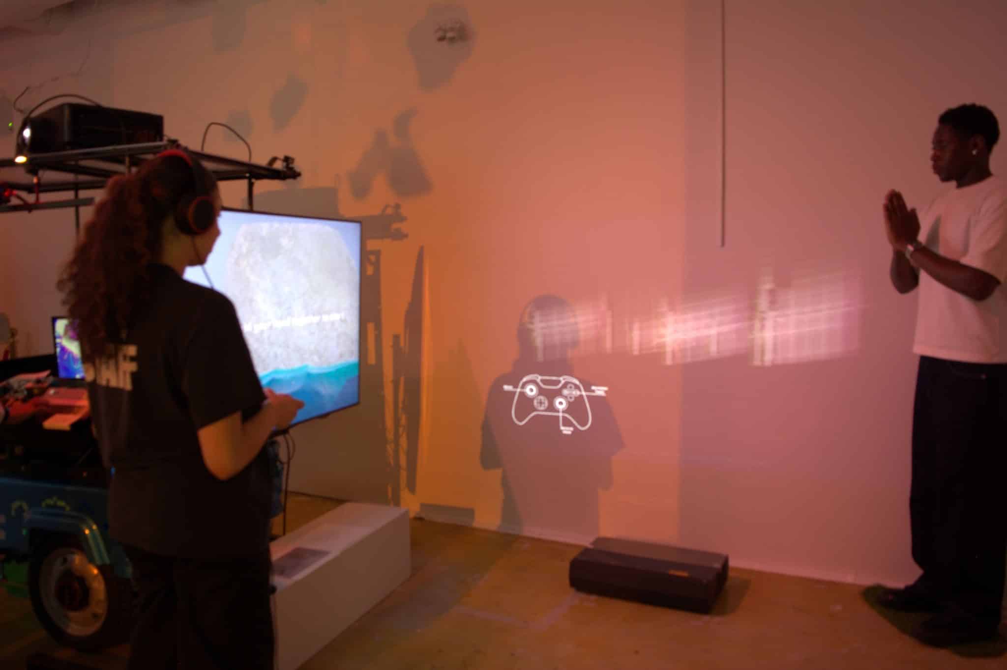 A visitor stands facing a screen in a dimly lit room while a second person watches, light projected across the wall.