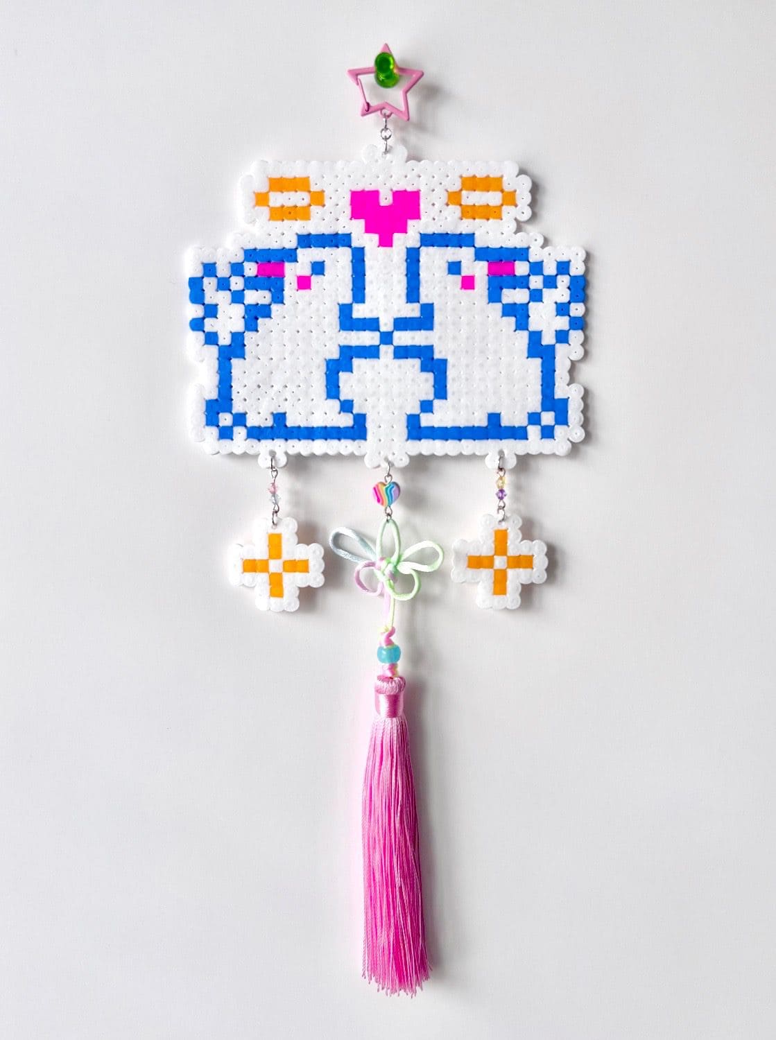 A perler bead panel of two blue rabbits facing each other, hung with beaded charms and a pink tassel.